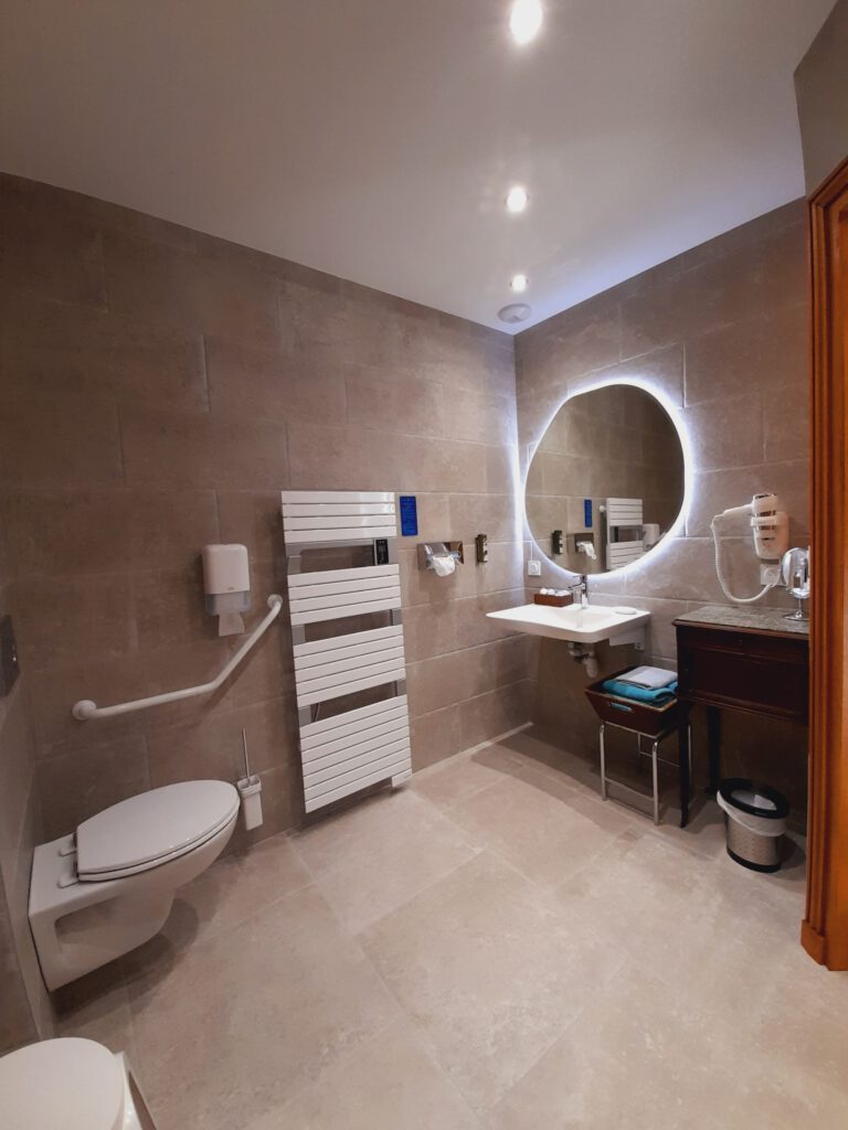 Bagno PMR moderno e spazioso con attrezzatura accessibile, piastrelle leggere e illuminazione leggera.