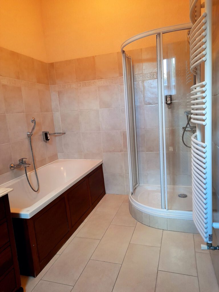 Amplio baño con bañera y ducha, decorada con baldosas beige y muebles de madera. Iluminación cálida.